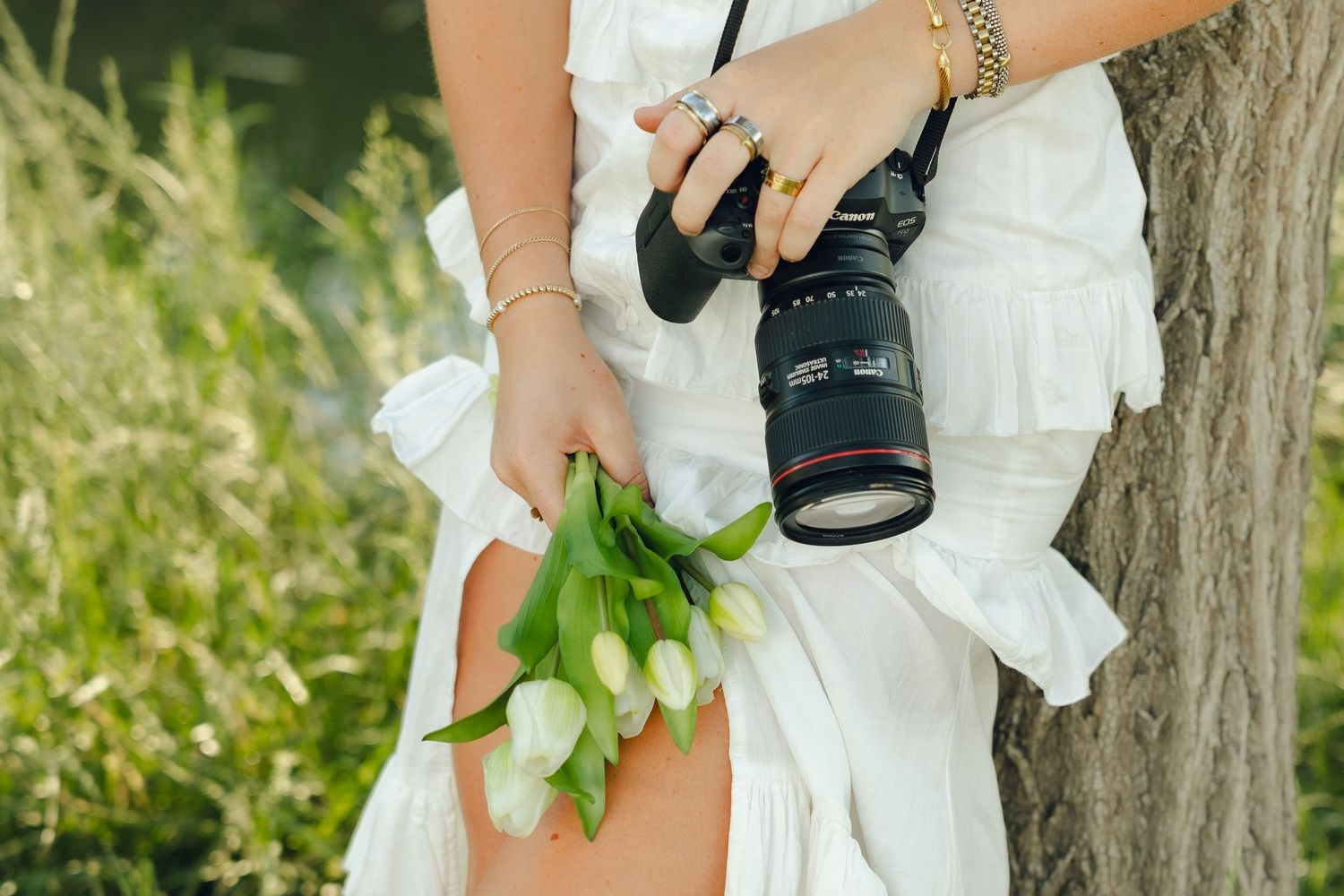 A photographer in a white dress holds a professional camera and fresh white tulips while standing near a tree outdoors.