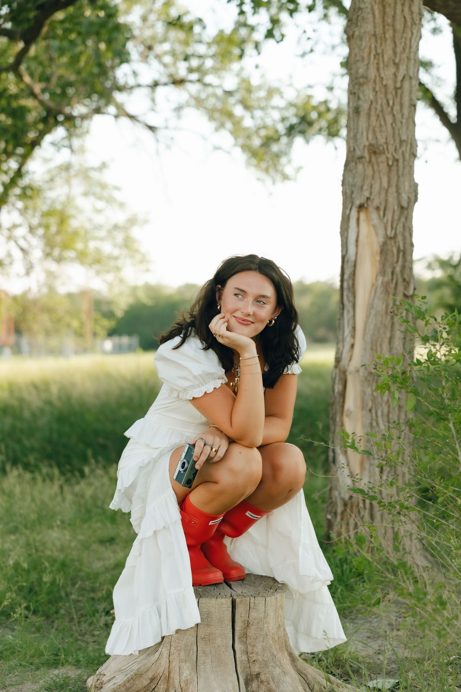 Someone in a flowing white dress and red rain boots sits casually on a tree stump in a summer field.
