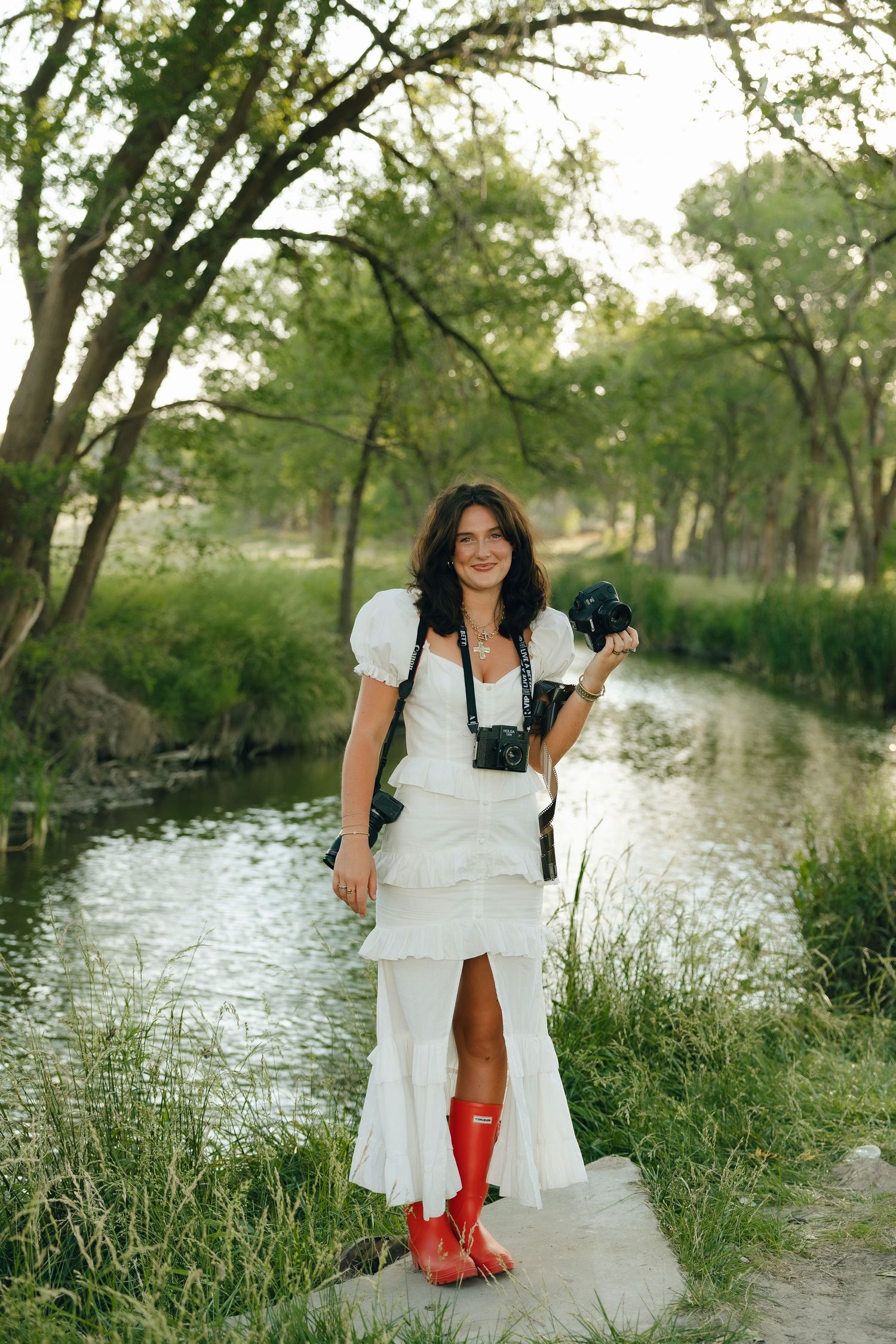 A photographer in a white ruffled dress and red rain boots poses along a scenic creek with trees in the background.