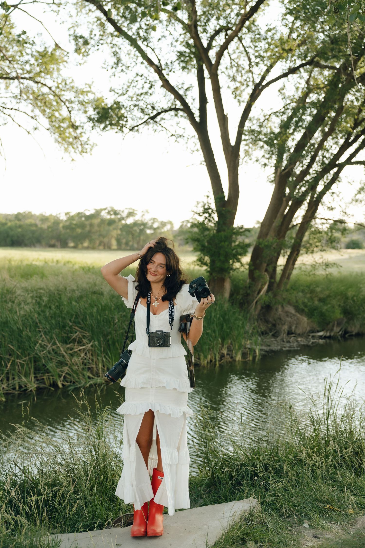 A creative portrait by a stream features a white ruffle dress, red rain boots and camera gear in nature.