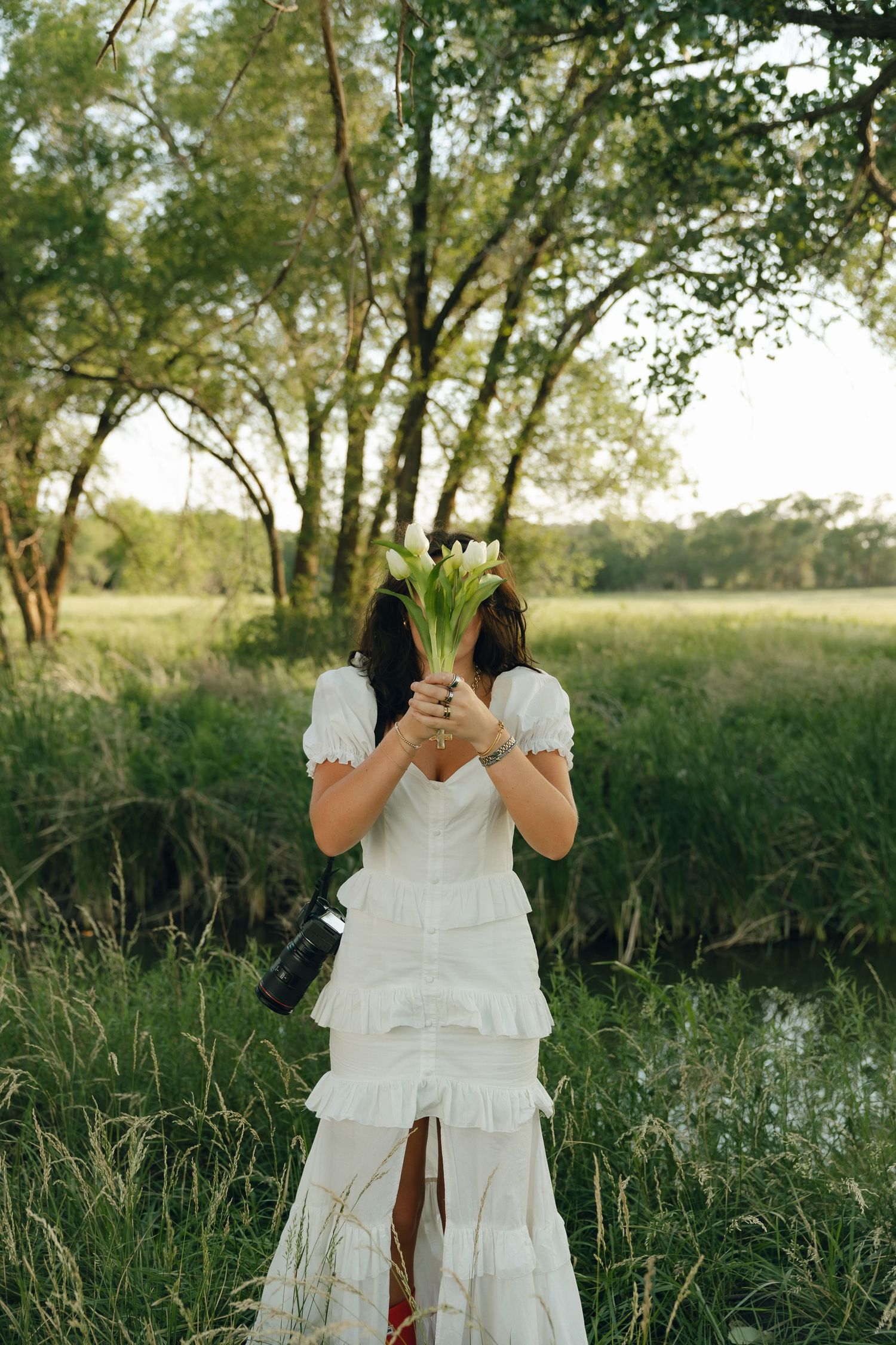 Photographer in a white tiered dress holds flowers while standing in a golden-lit meadow with trees.