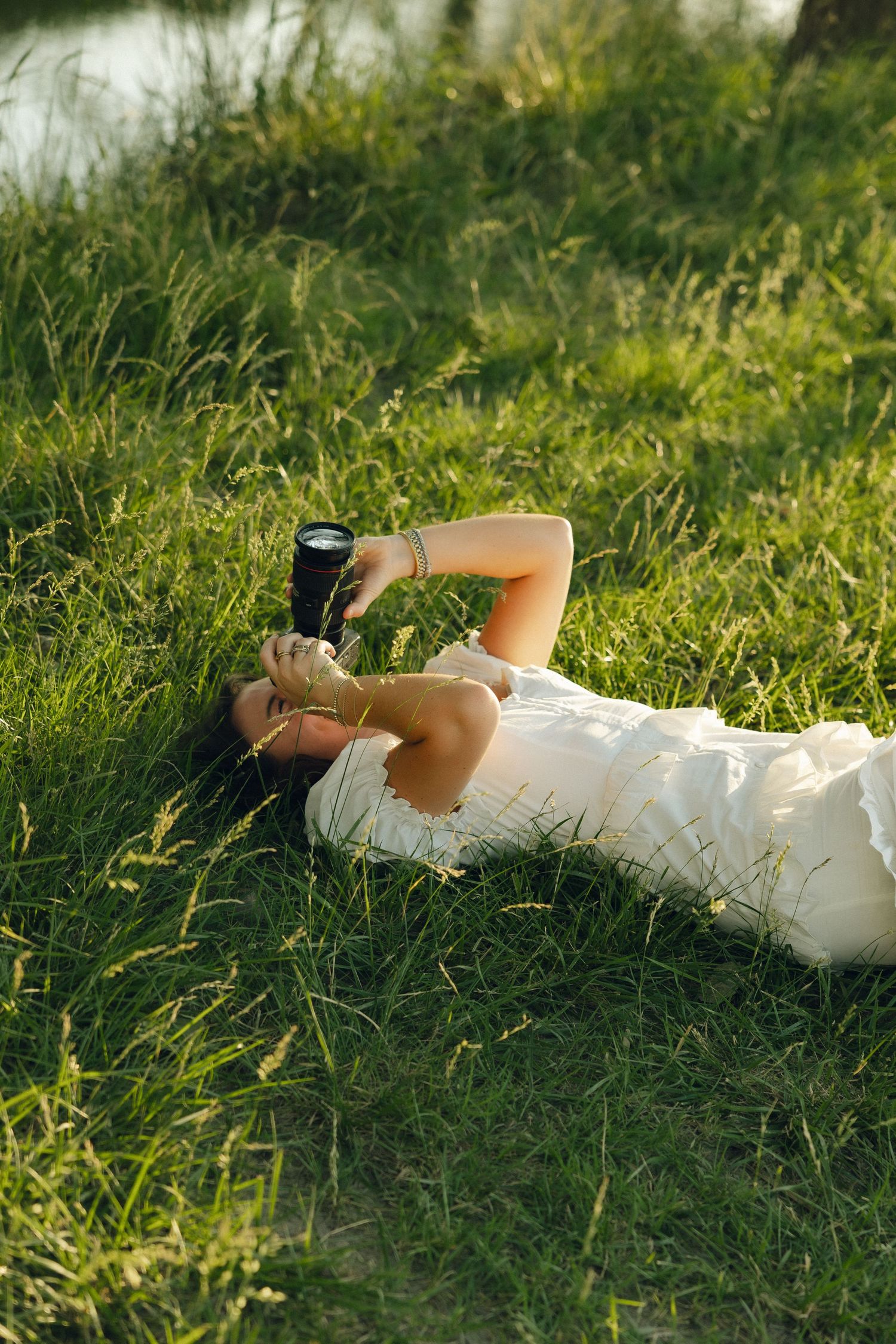 Photographer in white dress lies on grass while taking photos during golden hour.