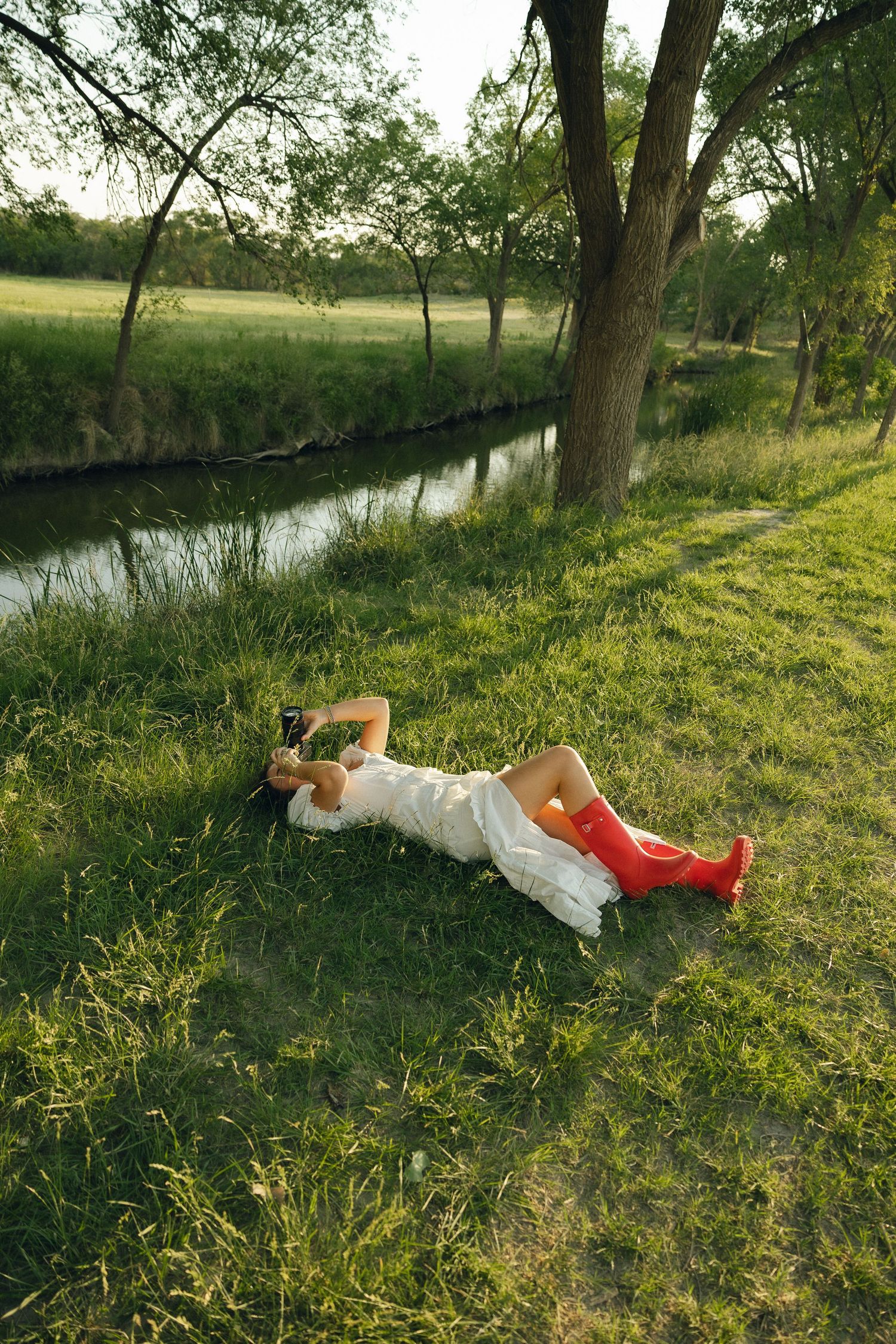 Person in white dress and red boots lying in grass by a peaceful stream on a sunny afternoon.