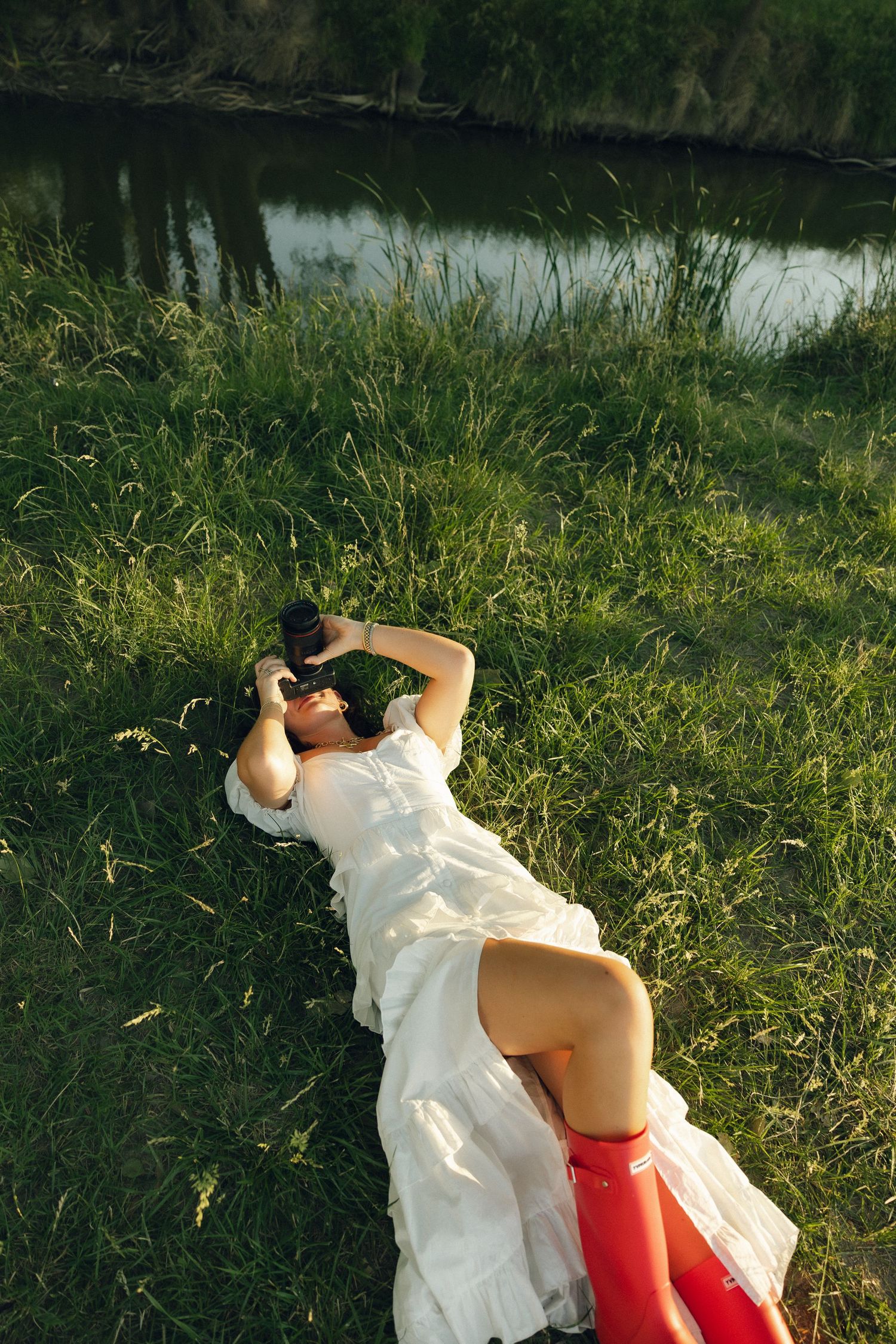 A person in white dress and red boots lies on grass by a riverbank while taking photos with a camera.