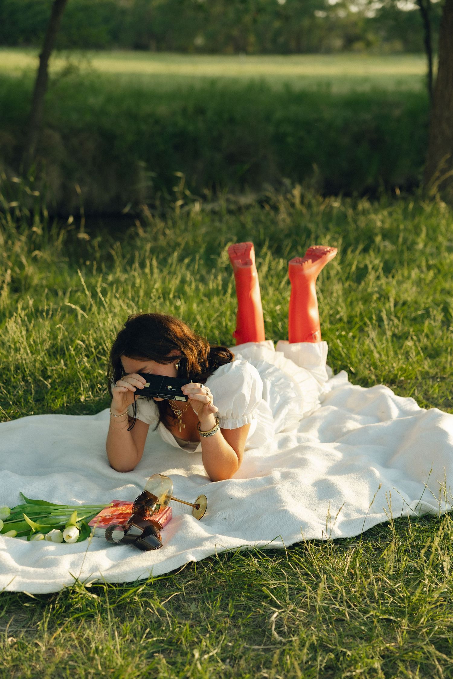 Photographer wearing red boots lies on white blanket taking photos in sunny meadow.