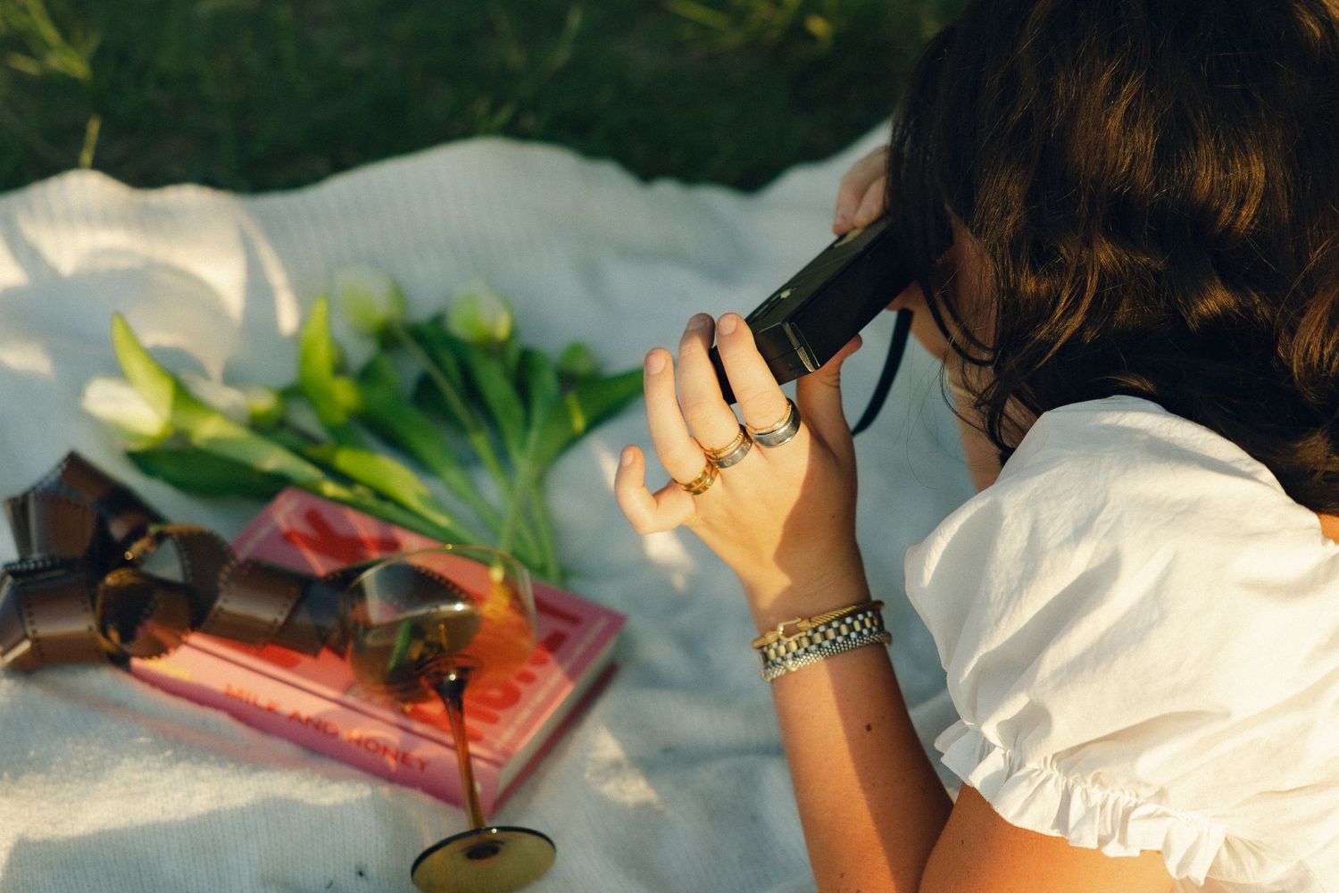 Someone photographing a picnic scene with flowers and books laid out on a white blanket in sunlit outdoor setting.