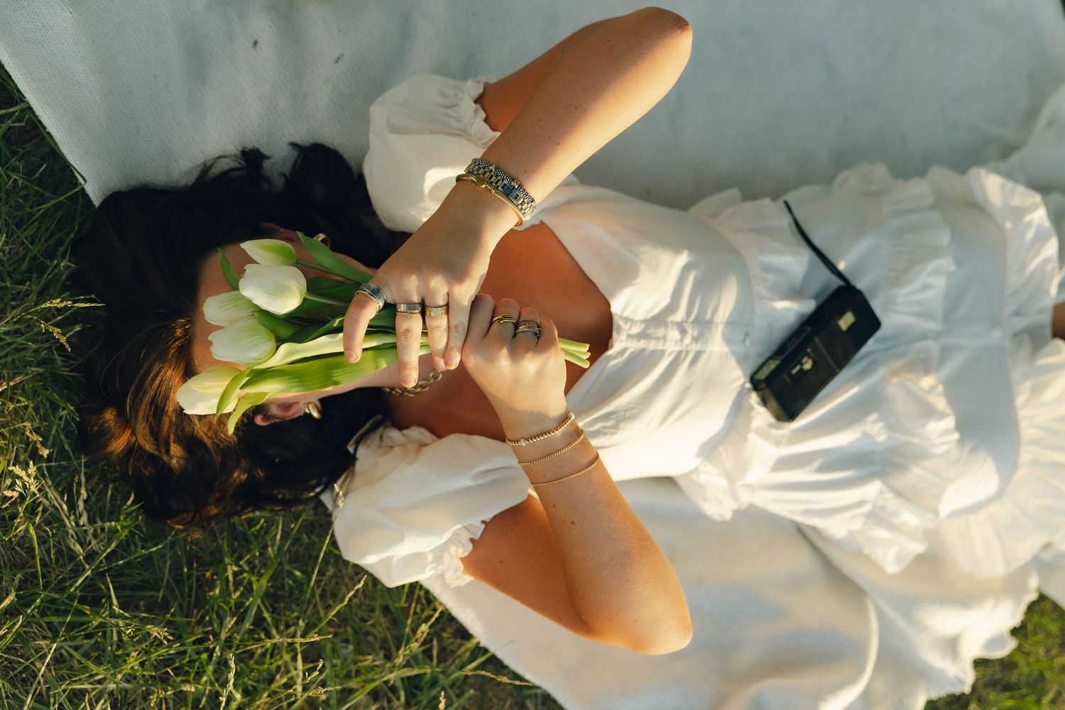 Someone in white dress lying on grass holding vintage camera on summer day.