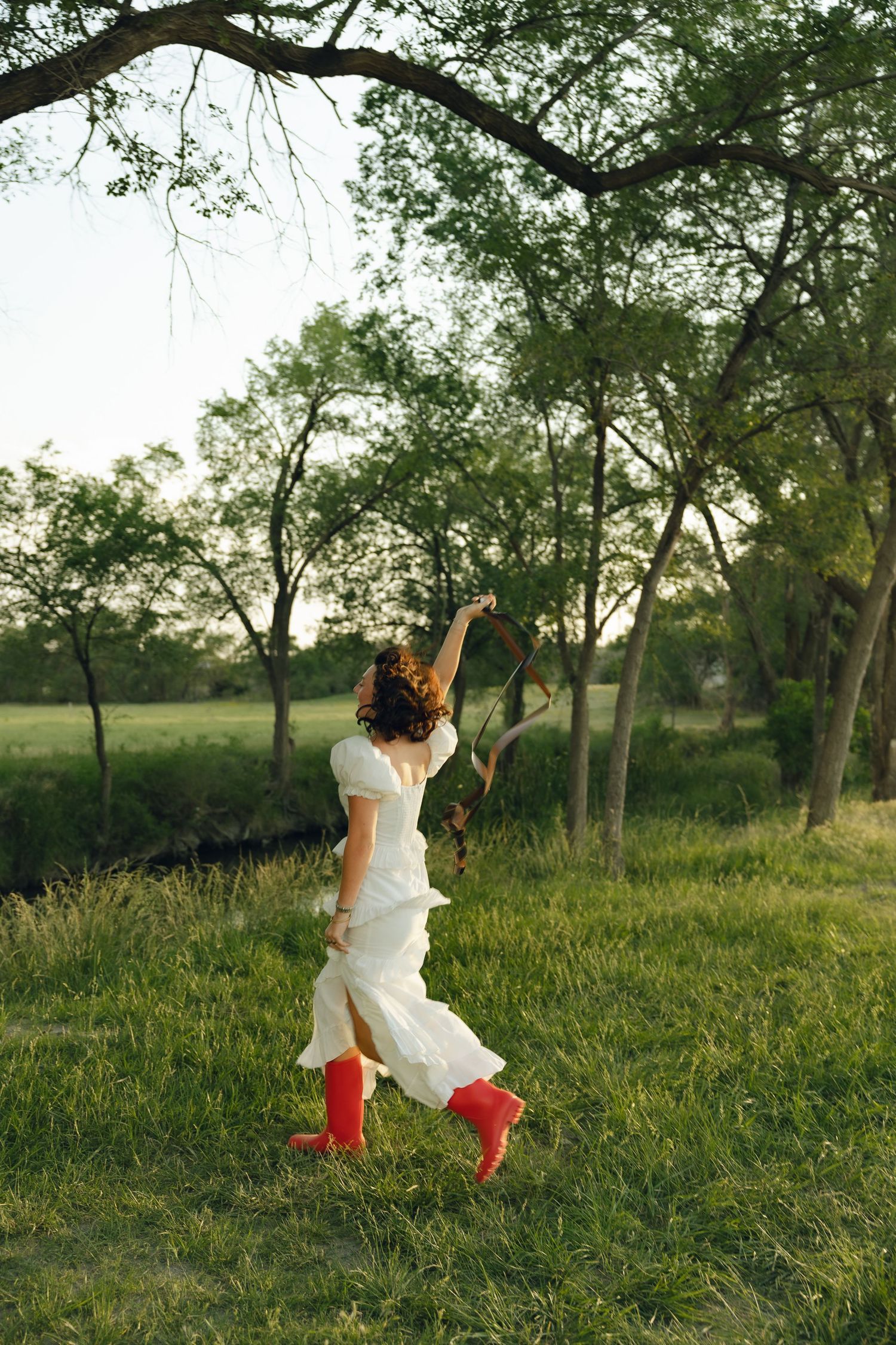 Figure in flowing white dress and red boots dances joyfully in a sunlit field with trees.