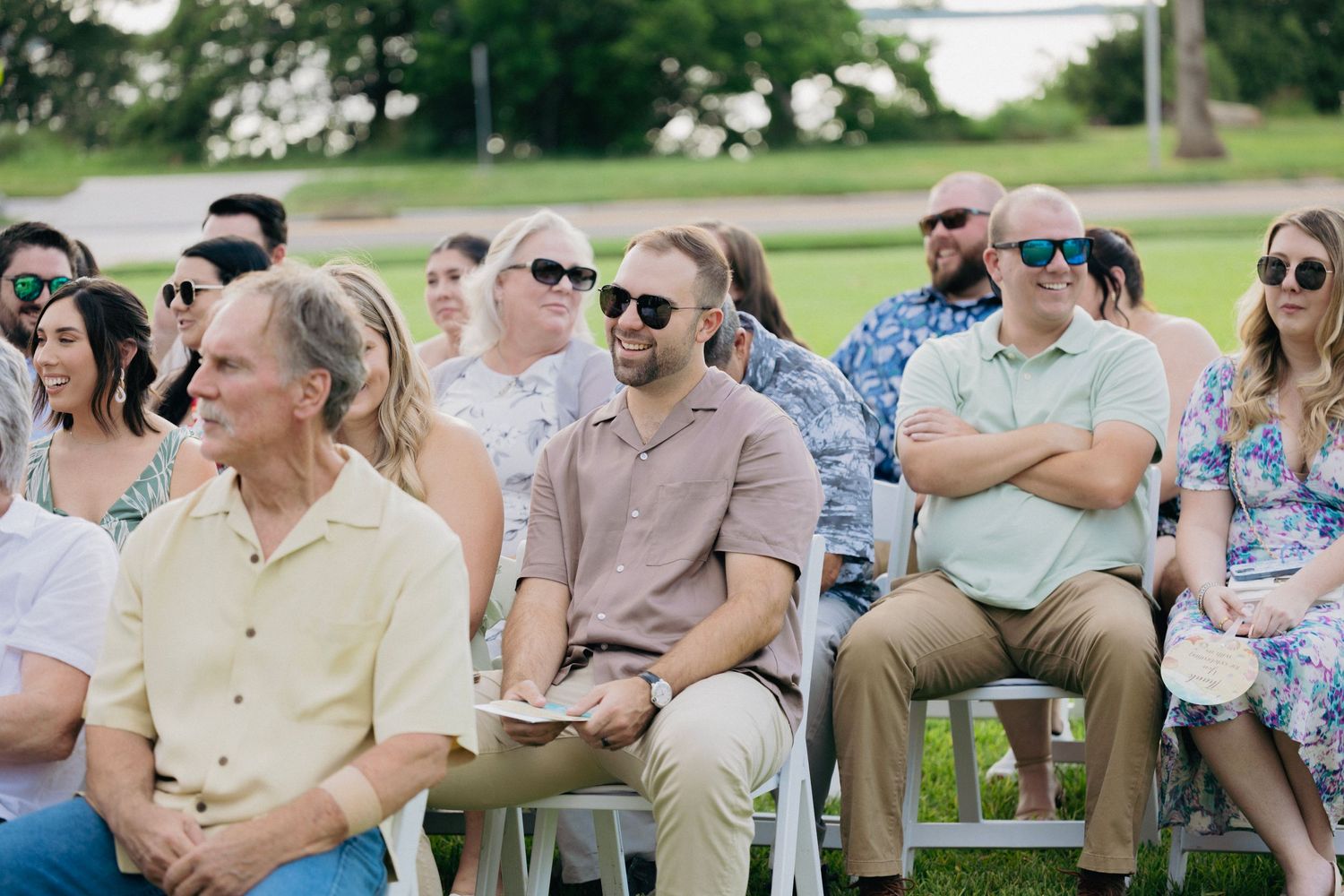 Wedding guests seated outdoors in white chairs watching a sunny daytime ceremony.