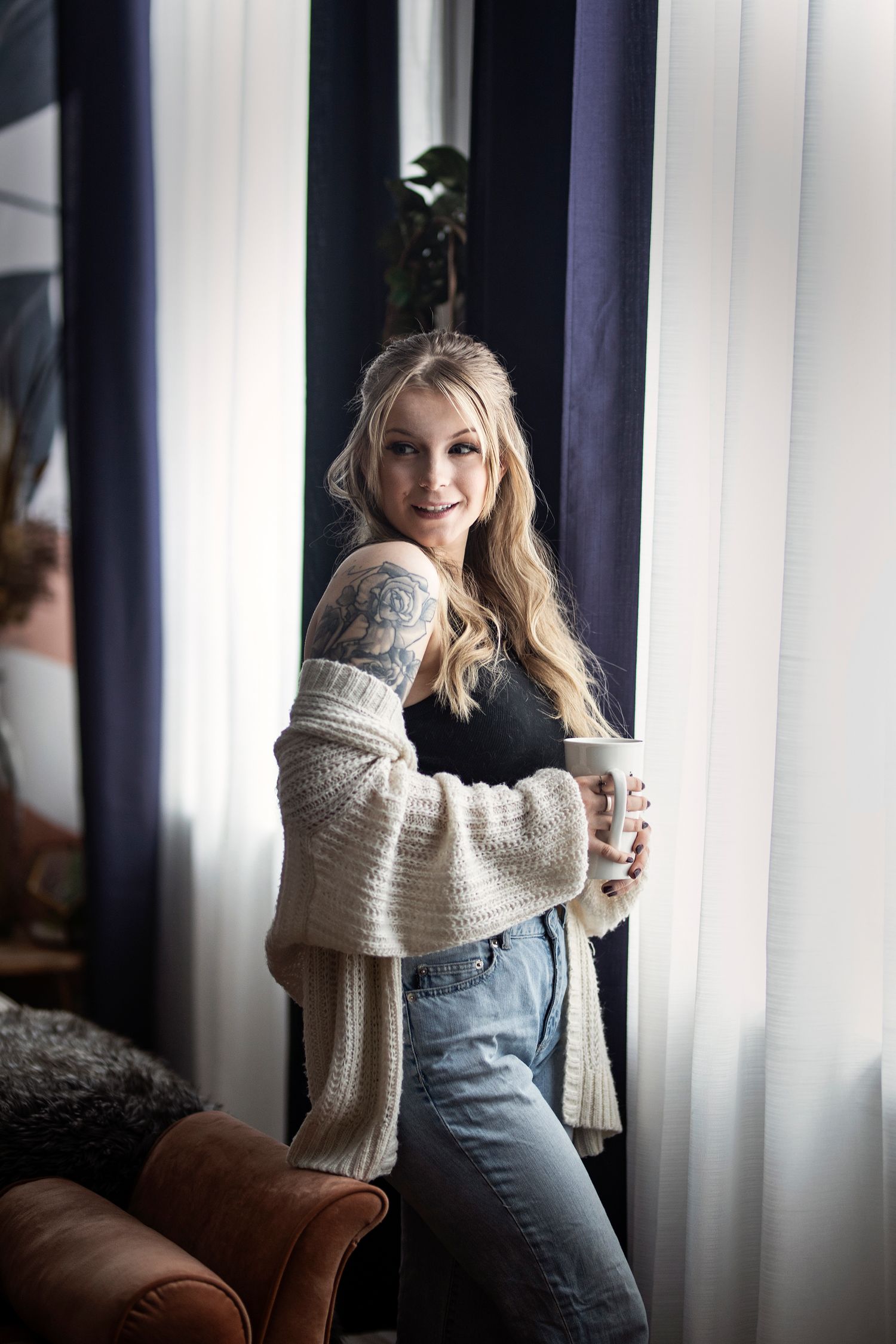 Woman in cozy sweater and jeans poses by window with soft natural lighting and warm interior decor.