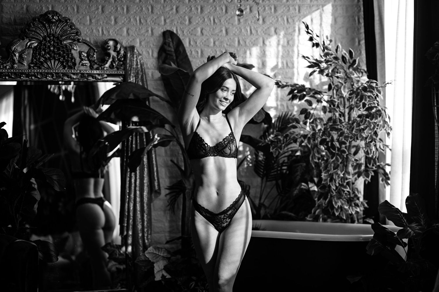 Black and white boudoir photograph with vintage-style mirror and natural window lighting.