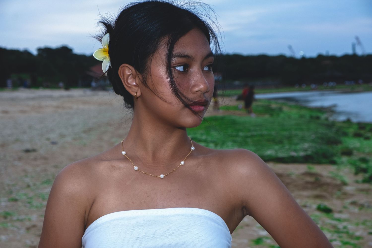 Woman in white dress with flower in hair poses against beach sunset backdrop.