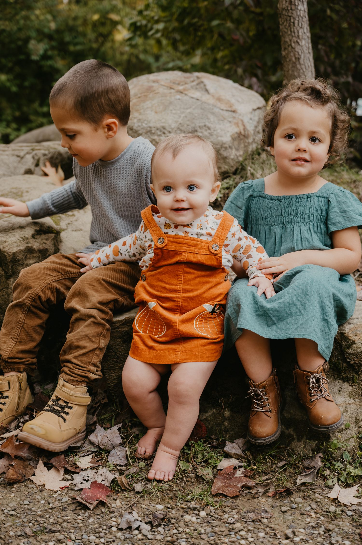 Three young siblings sitting on rocks outdoors, with baby in orange overalls in the center.