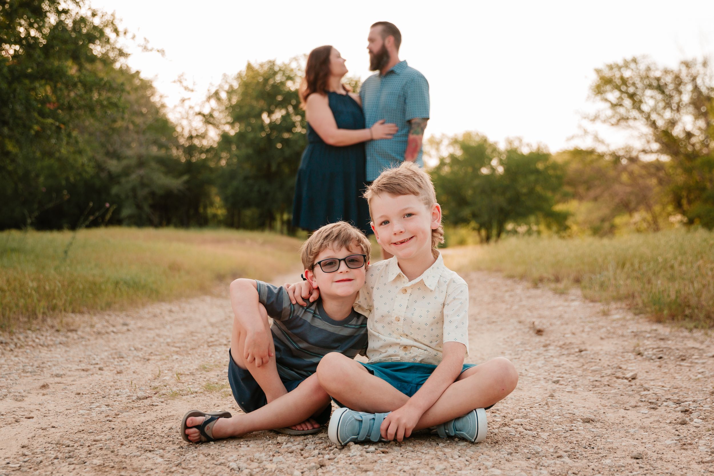 Two children sit on a dirt path while adults share a moment in the background during golden hour.