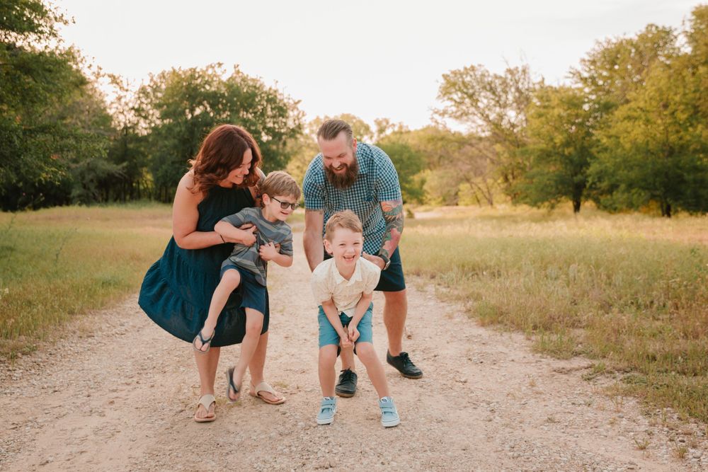 A playful family moment captured on a dirt path surrounded by green fields at sunset.