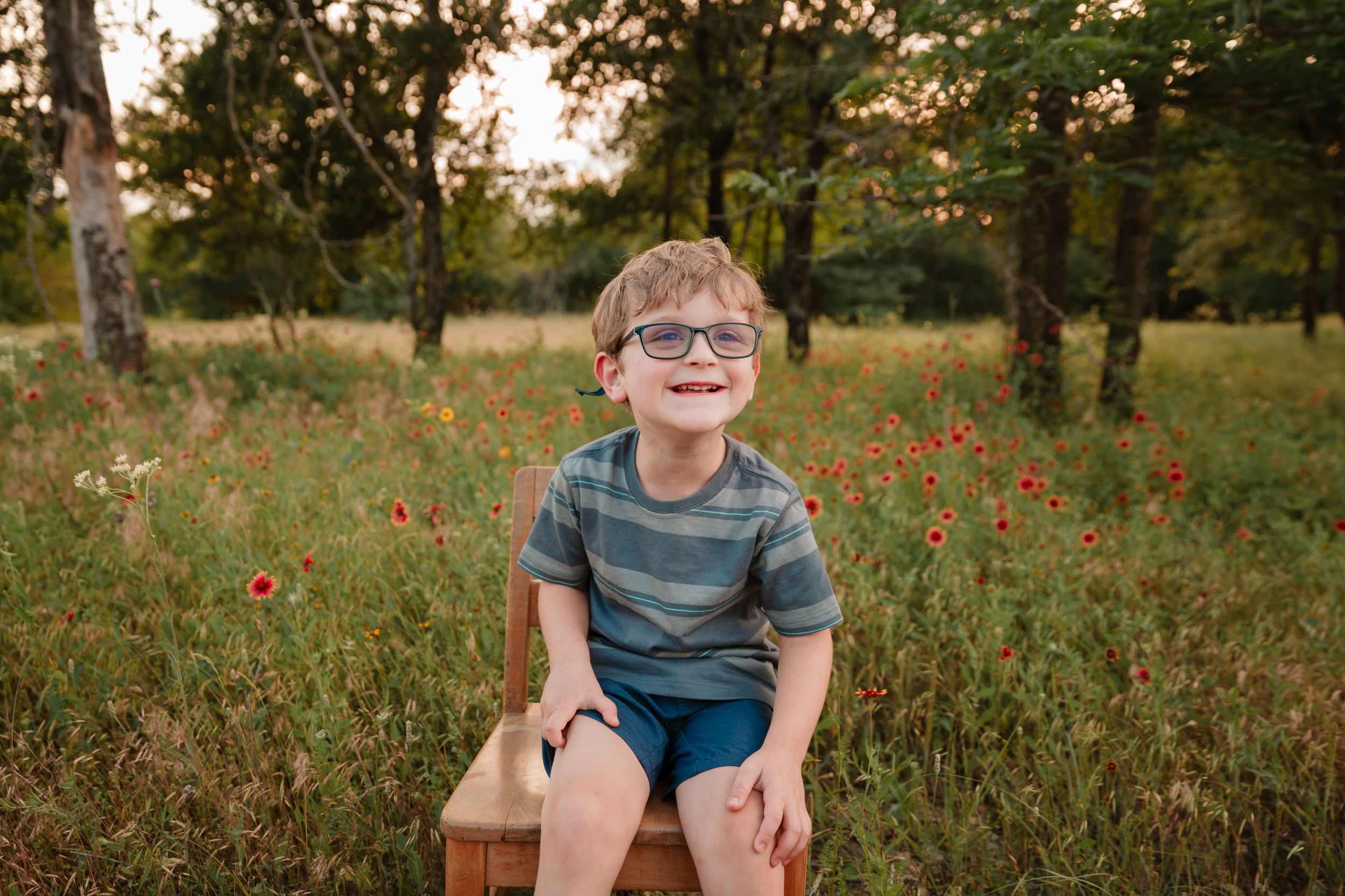 A seated little boy poses on a wooden bench amid wildflowers and trees.
