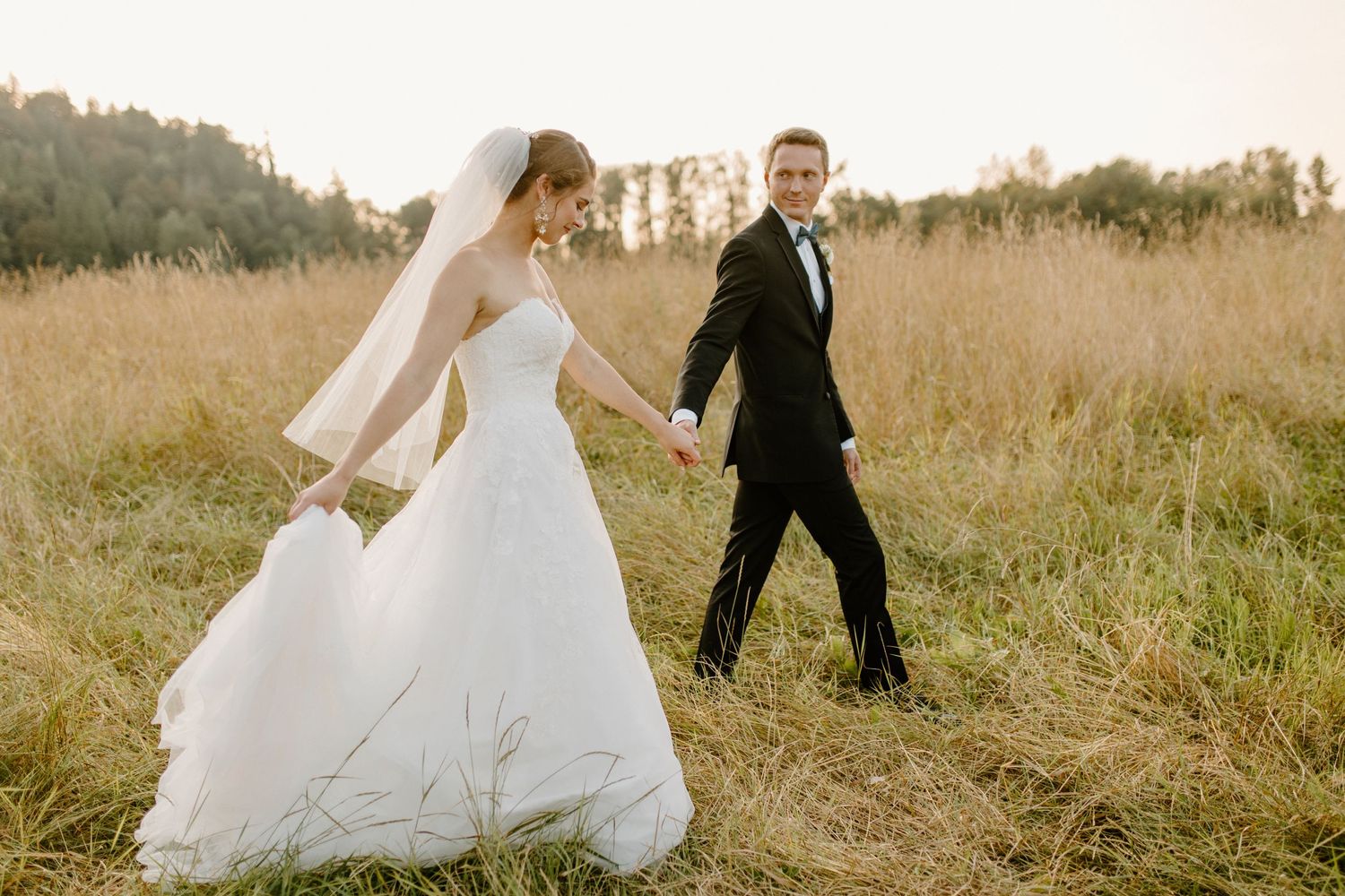 A couple walks hand in hand through a golden field during their sunset wedding photoshoot.