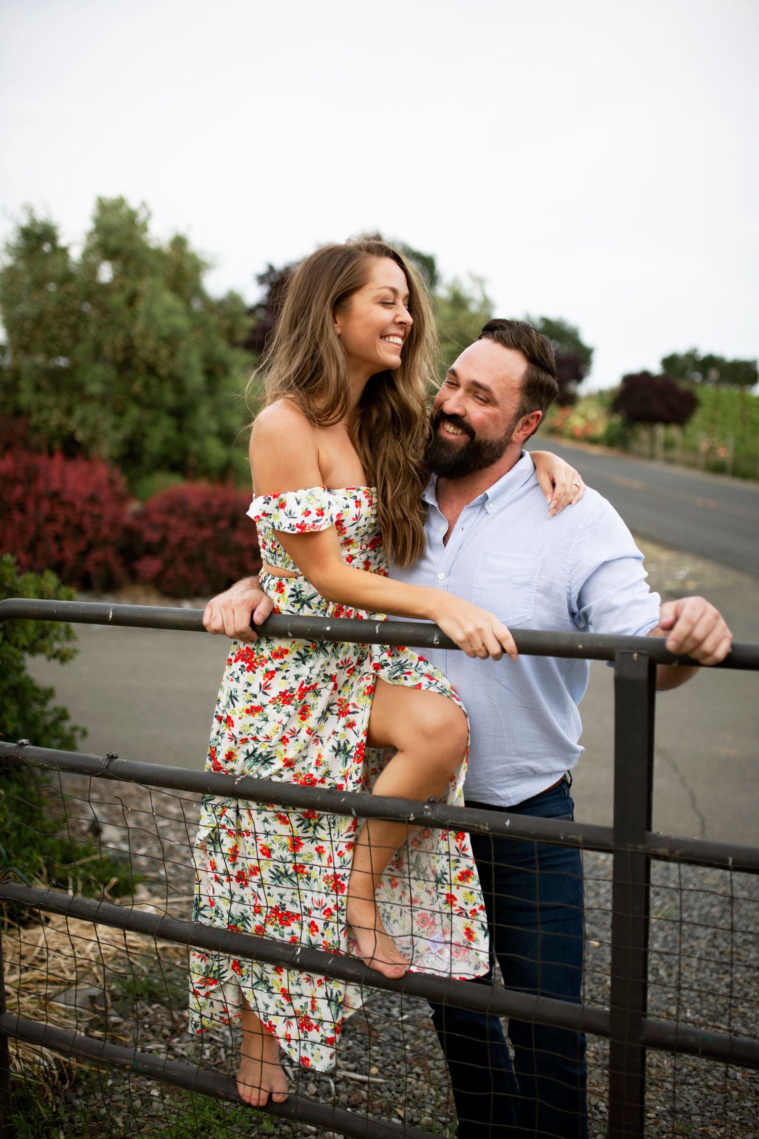 Couple leaning on a rustic gate along a countryside road in Healdsburg, she in a floral dress laughing with greenery in the background.