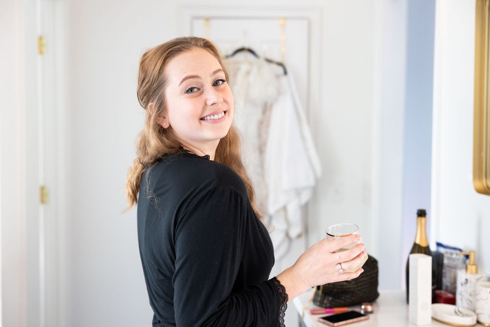A person in black attire smiling while getting ready in a room with a dress visible in background.