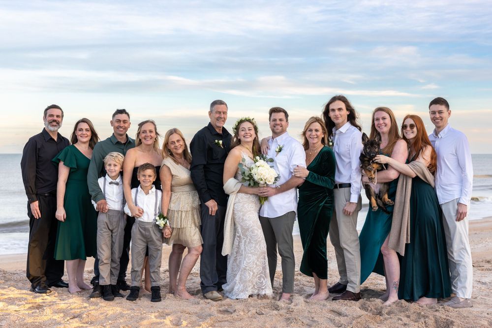 Large group photo of wedding party and family members gathered on beach at sunset.