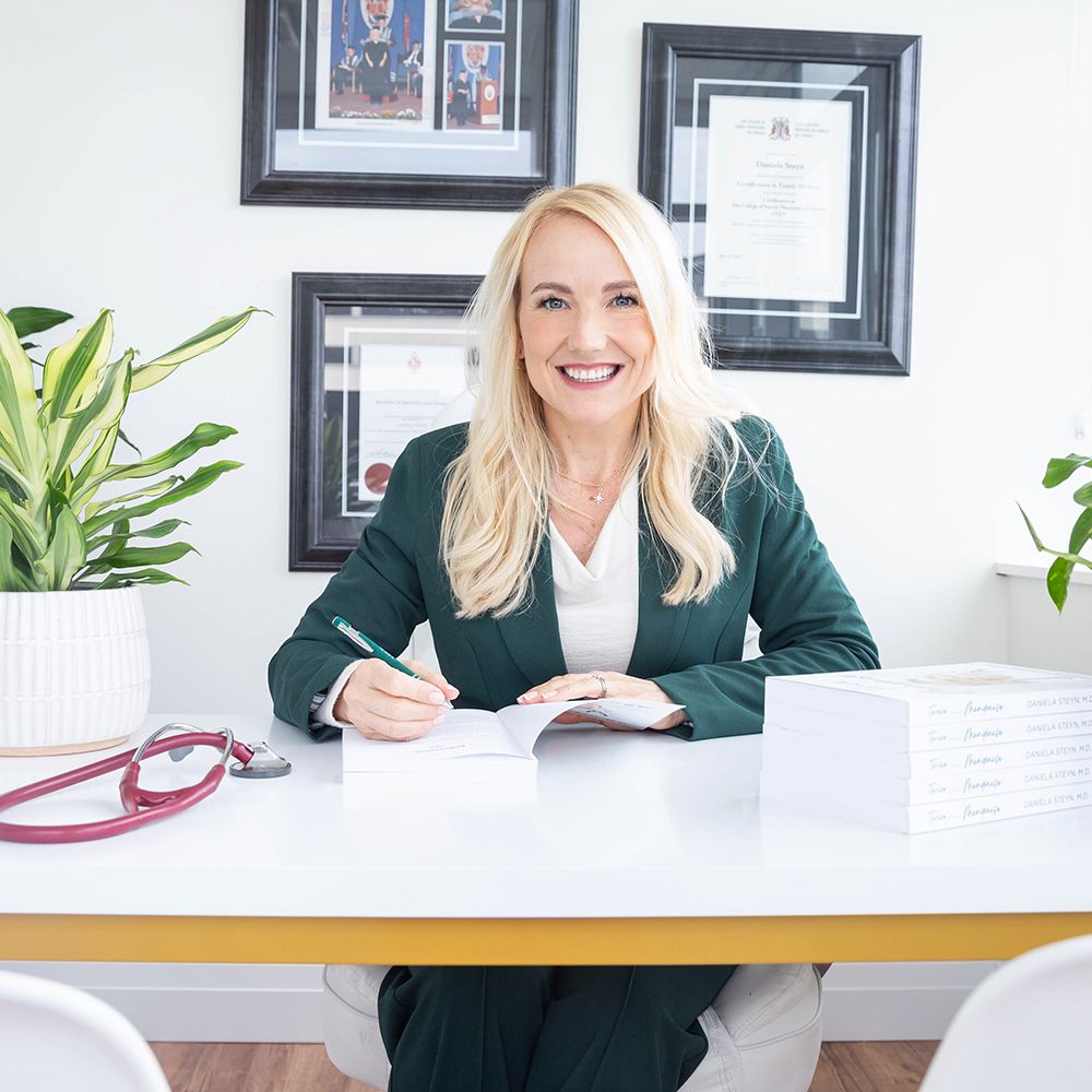 Professional in green blazer sits at modern white desk with framed certificates on wall and houseplant.