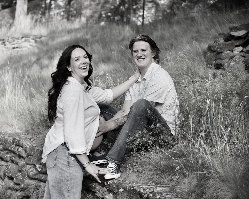 Black and white photo of a laughing couple sitting outdoors on rocks surrounded by tall grass and wildflowers.