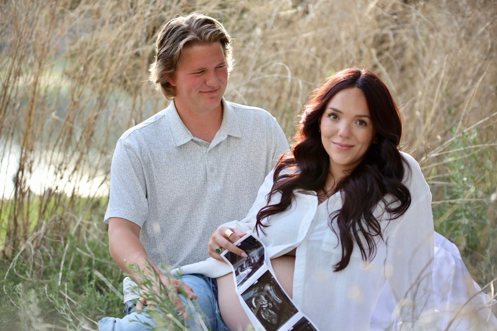 Couple sitting outdoors holding ultrasound photos, announcing pregnancy in a natural field setting.
