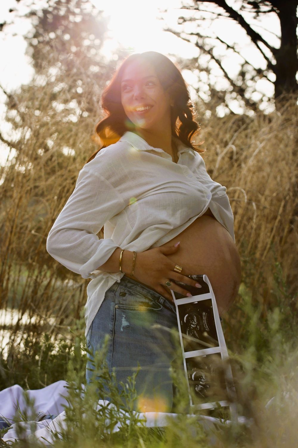Pregnant woman outdoors at golden hour, holding ultrasound photo against bare belly, wearing white shirt and denim skirt.
