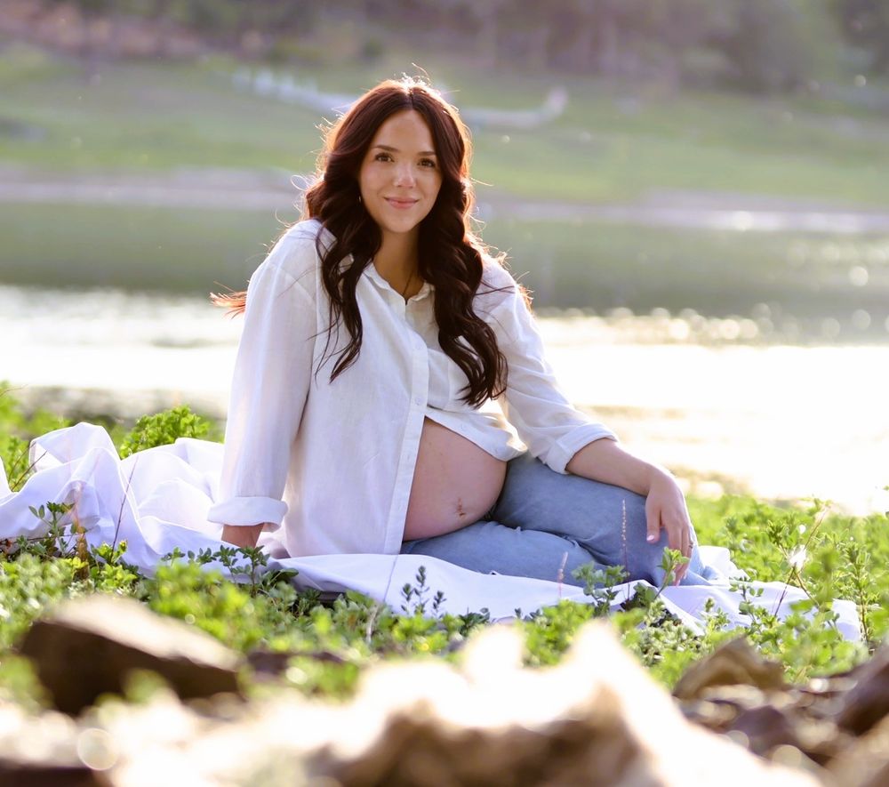 Pregnant woman in white shirt and jeans sits on blanket outdoors by water during golden hour maternity photo session.