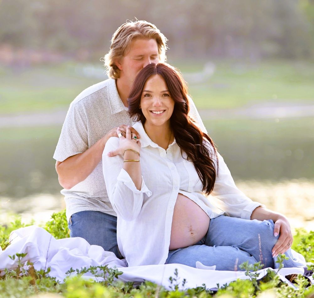 Happy couple sitting together outdoors showing pregnant belly during a sunny maternity photoshoot by a lake.