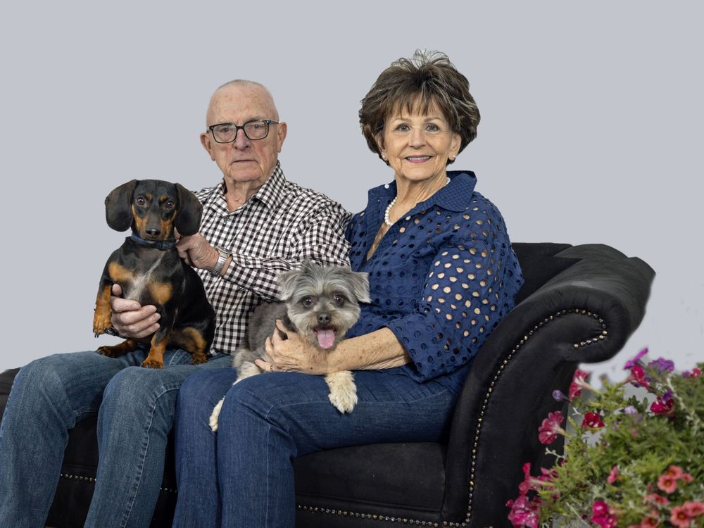 Couple sitting on sofa with small dogs in studio setting against grey background.