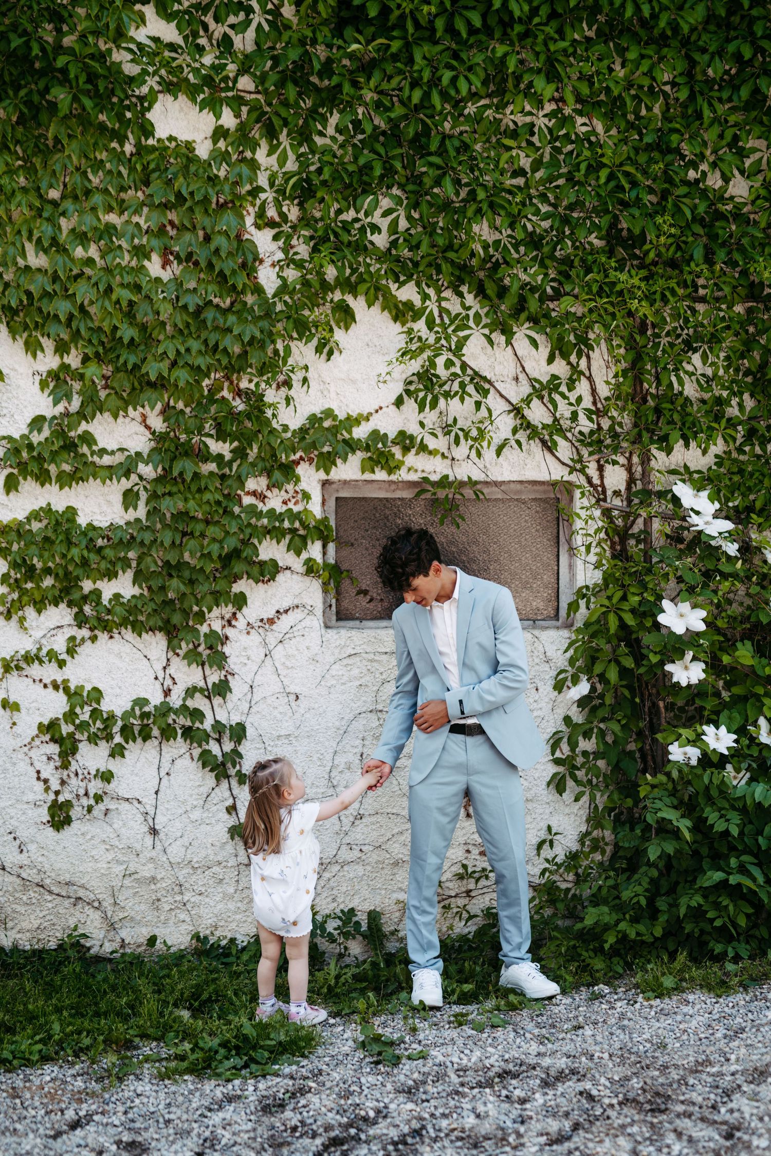 A touching outdoor moment between a small child and an adult in light blue suits against an ivy-covered wall.