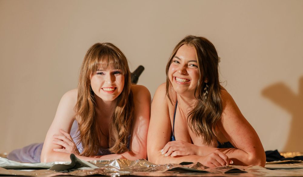 Two smiling young women in formal dresses lying on the floor posing together for a photo.