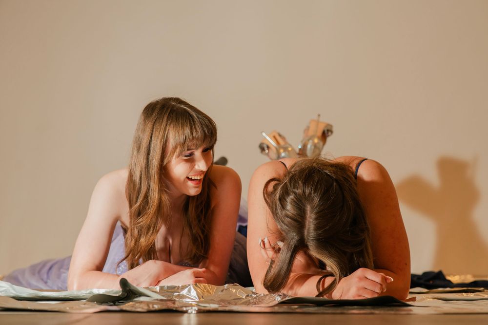 Two young women in formal gowns lying on the floor, one laughing while the other rests her head down.