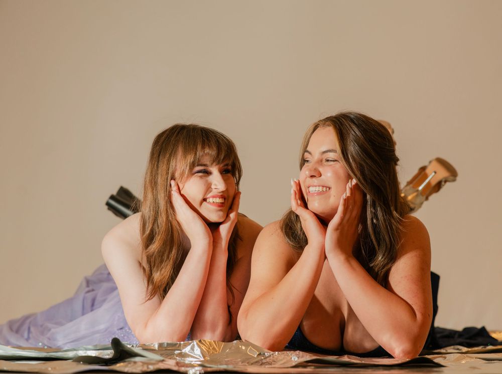 Two young women in formal dresses lying down smiling with hands on cheeks in a playful pose.