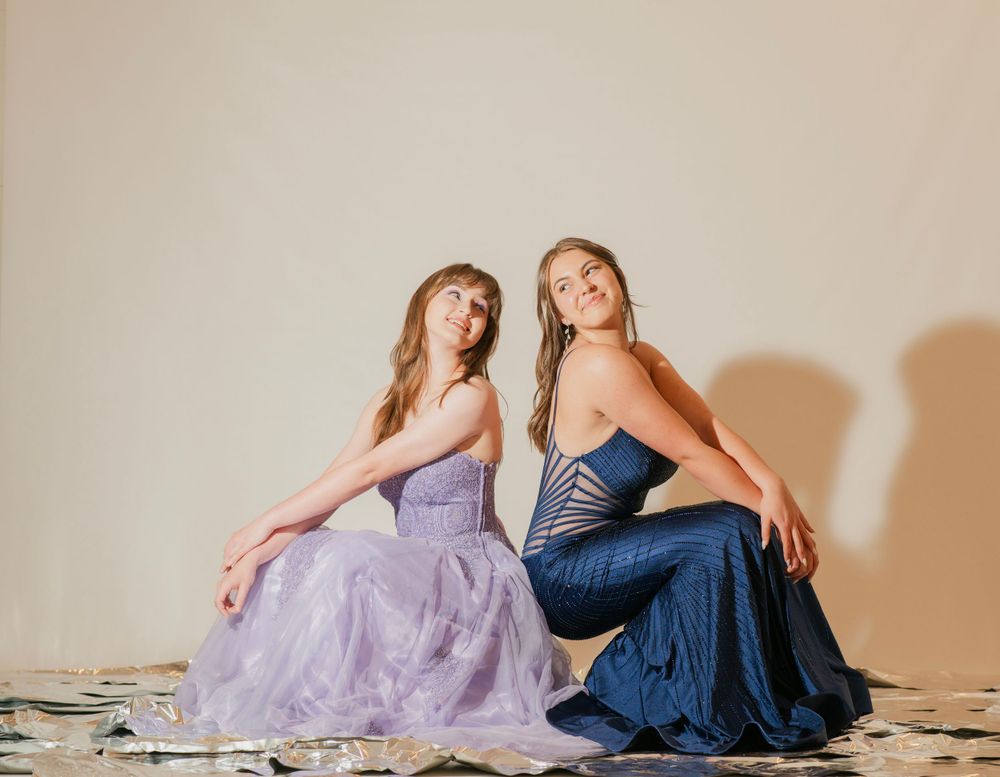 Two young women in elegant prom gowns, lavender and navy blue, sitting back to back on studio floor.