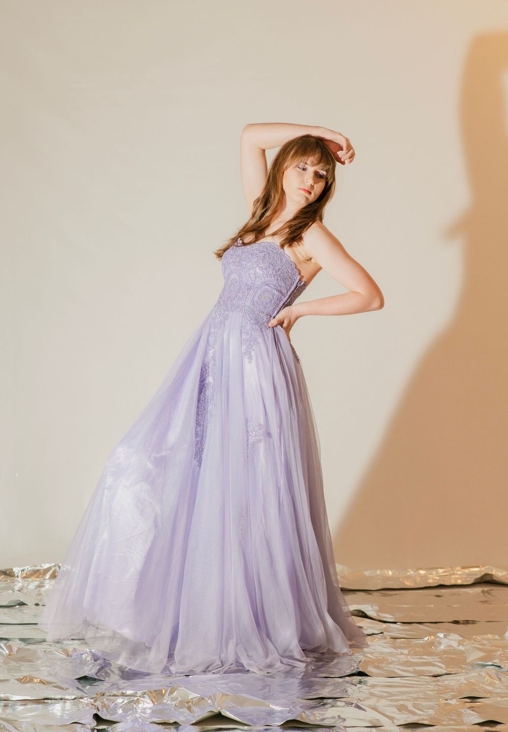 Young woman in lavender tulle A-line prom gown poses dramatically with arm raised in studio.