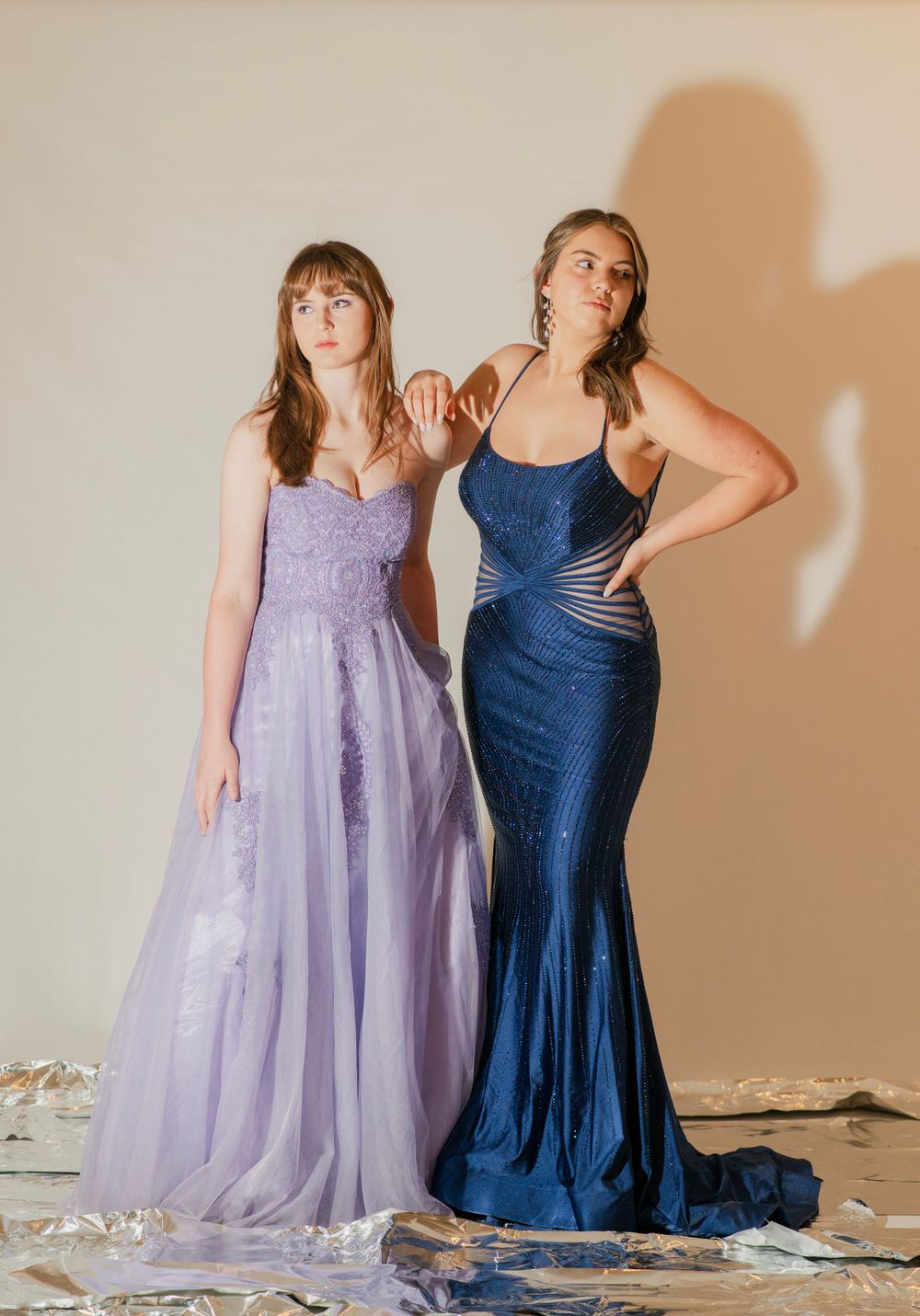 Two teens posing in formal gowns, one in lavender A-line and one in navy sequin mermaid dress.