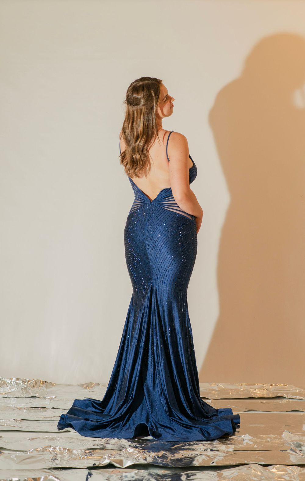 Woman in elegant navy blue sequin mermaid gown with open back and train, posing in studio.