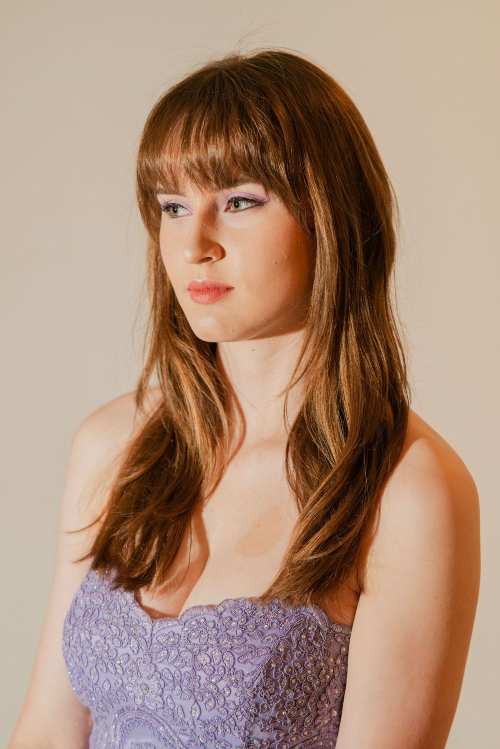 Young woman with long brown hair and bangs wearing strapless purple lace dress in studio portrait.