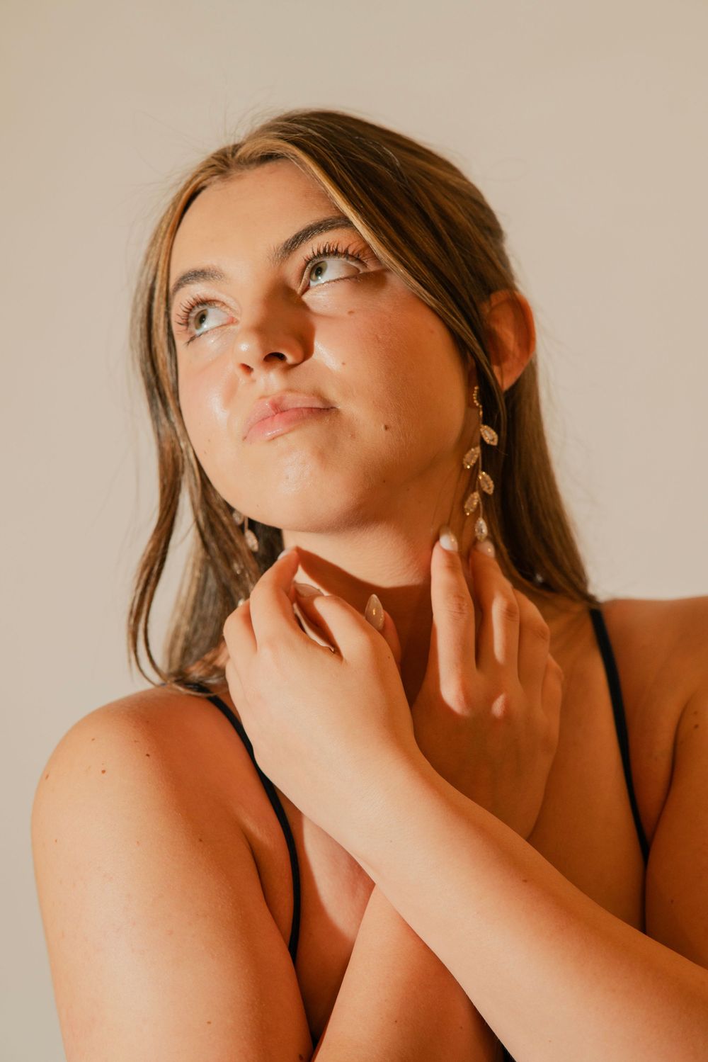 Young woman with long lashes wearing gold leaf earrings looking up, touching neck in studio.