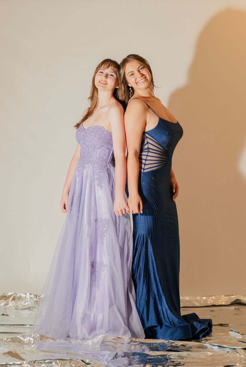 Two smiling young women in prom dresses, lavender tulle and navy sequin, posing back to back.
