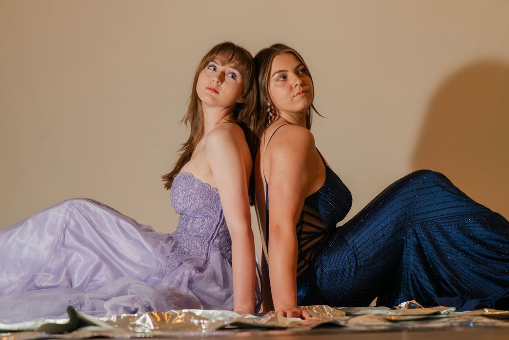Two young women in formal lavender and navy blue gowns sitting back-to-back on the floor posing.