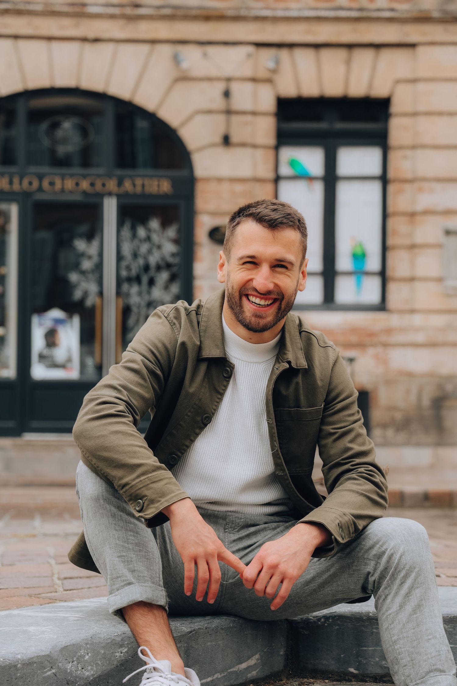 Smiling man in olive jacket and white turtleneck sitting outdoors in front of a chocolatier shop.