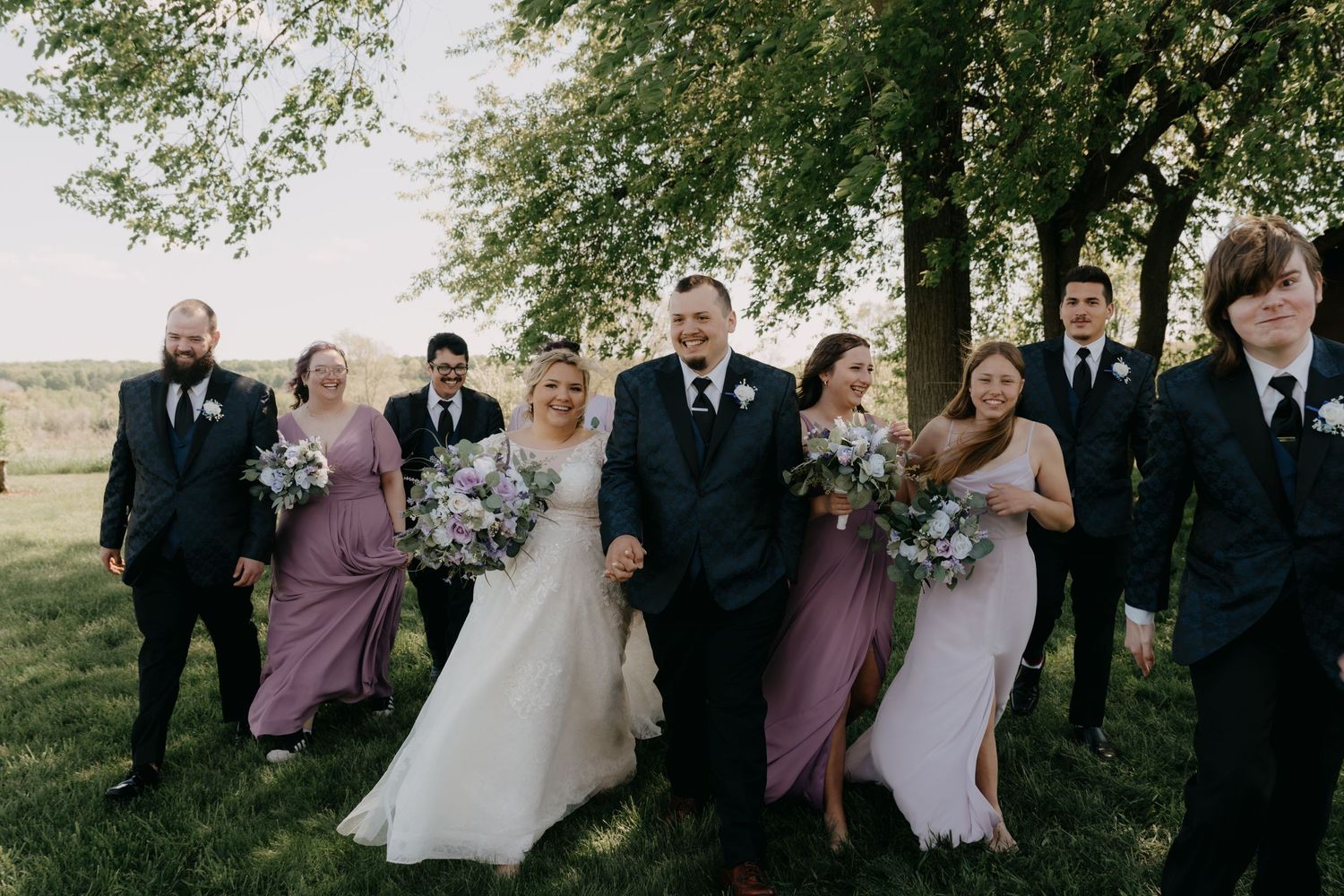 A wedding party poses together outdoors wearing purple bridesmaid dresses and black suits.