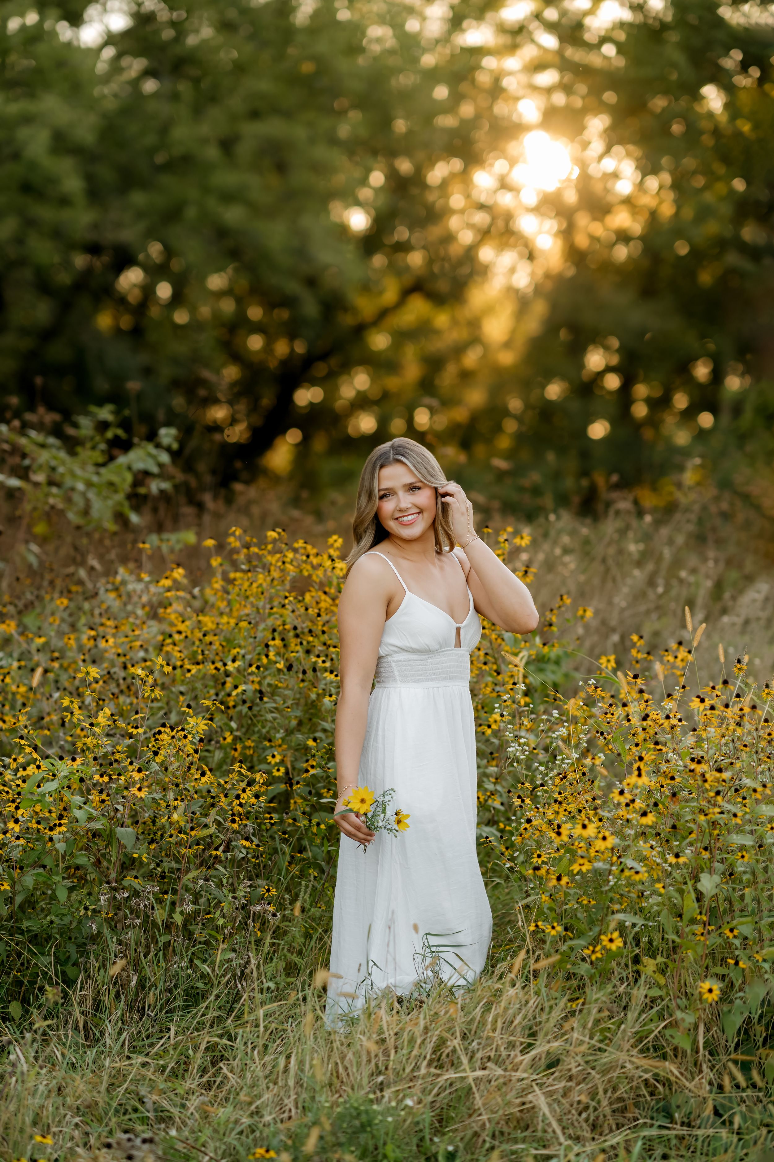 Smiling young woman in a white sundress holding yellow wildflowers in a field of black-eyed Susans at golden hour sunset.