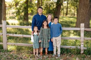 Family of five posing together outdoors in a sunny wooded park, standing in front of a rustic wooden fence surrounded by tall trees.