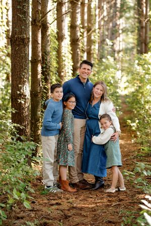 A smiling family of five in coordinated blue and neutral outfits poses together on a pine needle path in a sunlit forest.