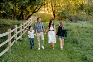 Family of four holding hands and walking along a wooden fence on a grassy path surrounded by trees in warm golden light.