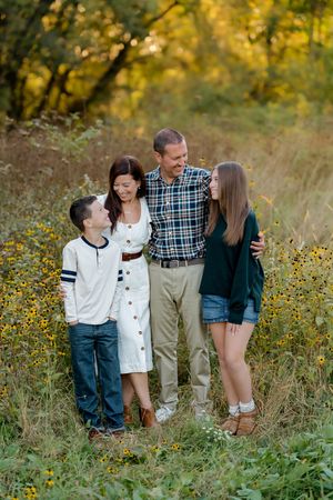 Family of four embracing and smiling in a golden field of yellow wildflowers during a sunset outdoor portrait session.
